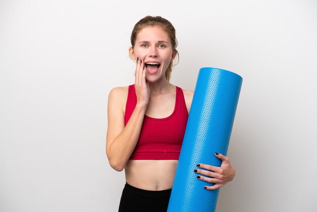 Giovane donna inglese sportiva che va a lezioni di yoga mentre tiene un tappetino isolato su sfondo bianco con espressione facciale sorpresa e scioccata