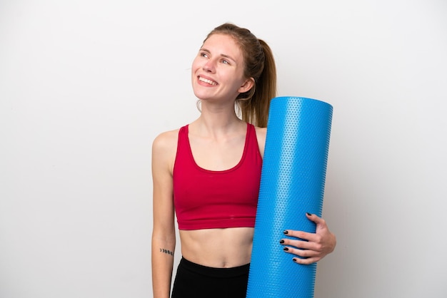 Giovane donna inglese sportiva che va a lezioni di yoga mentre tiene un tappetino isolato su sfondo bianco pensando a un'idea mentre guarda in alto