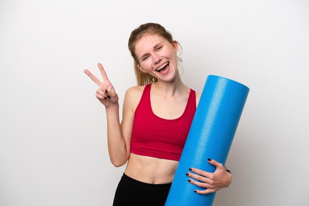 Giovane donna inglese sportiva che va a lezioni di yoga mentre tiene un tappetino isolato su sfondo bianco sorridente e mostra il segno della vittoria