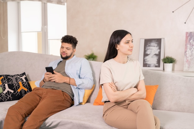Giovane donna insoddisfatta in abbigliamento casual che incrocia le braccia per il petto mentre è seduta sul divano contro il marito che scorre sullo smartphone