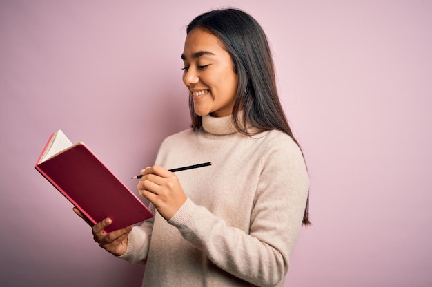 Giovane donna intelligente asiatica che tiene il diario del libro in piedi su sfondo rosa isolato con una faccia felice in piedi e sorridente con un sorriso fiducioso che mostra i denti