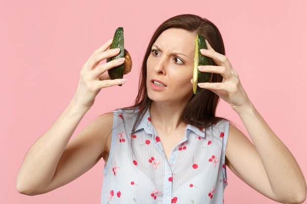 Giovane donna interessata in abiti estivi con due metà di avocado fresco maturo isolato su sfondo rosa pastello in studio. Lo stile di vita vivido delle persone si rilassa il concetto di vacanza. Mock up spazio di copia.