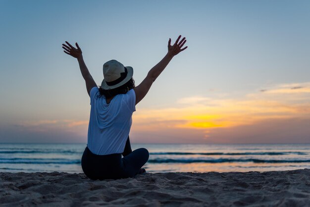 Giovane donna irriconoscibile con il cappello che alza le braccia eccitata per il tramonto sulla spiaggia