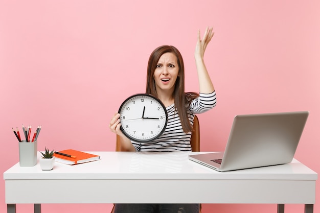 Giovane donna irritata che allarga le mani tenendo la sveglia seduta, lavora in ufficio con un computer portatile