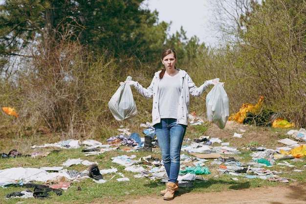 Giovane donna irritata e sconvolta in abiti casual che pulisce tenendo in mano i sacchetti della spazzatura e allargando le mani in un parco disseminato di rifiuti