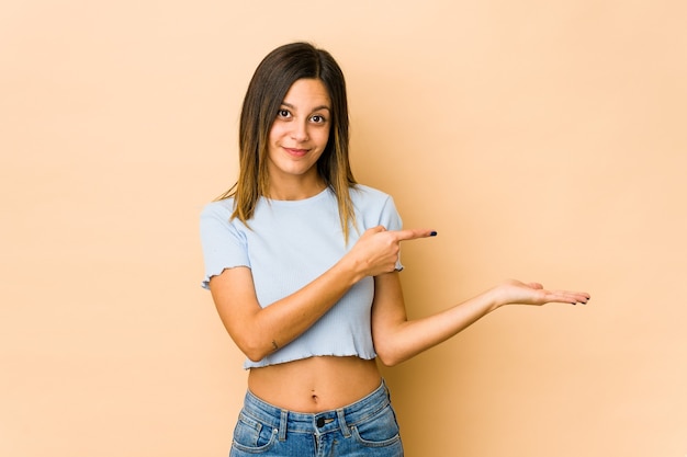 Giovane donna isolata su beige eccitato che tiene uno spazio della copia sul palmo.