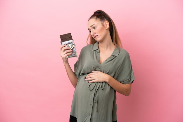 Giovane donna isolata su sfondo rosa incinta e in possesso di cioccolato con fame