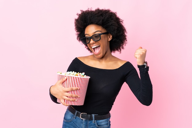 Giovane donna isolata sul rosa con gli occhiali 3d e che tiene un grande secchio di popcorn