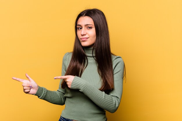 Giovane donna isolata sulla parete gialla scioccata che punta con il dito indice a uno spazio di copia.