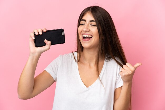 Giovane donna isolata sulla parete rosa utilizzando il telefono cellulare e cantando