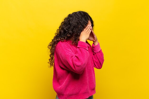 Giovane donna ispanica che copre gli occhi con le mani con uno sguardo triste e frustrato di disperazione, pianto, vista laterale