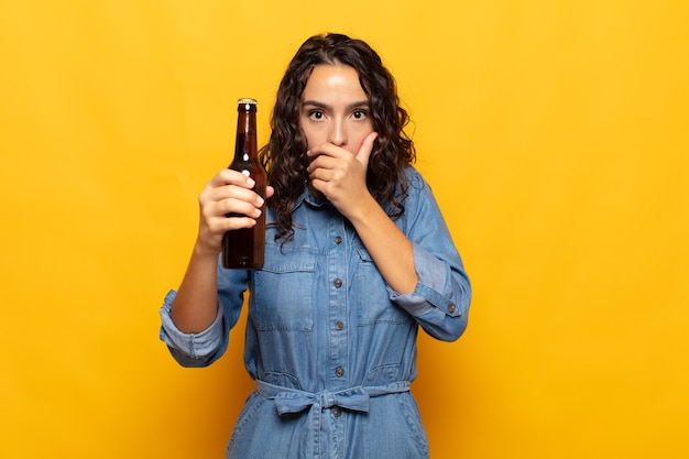 Giovane donna ispanica che copre la bocca con le mani con un'espressione scioccata e sorpresa, mantenendo un segreto o dicendo oops