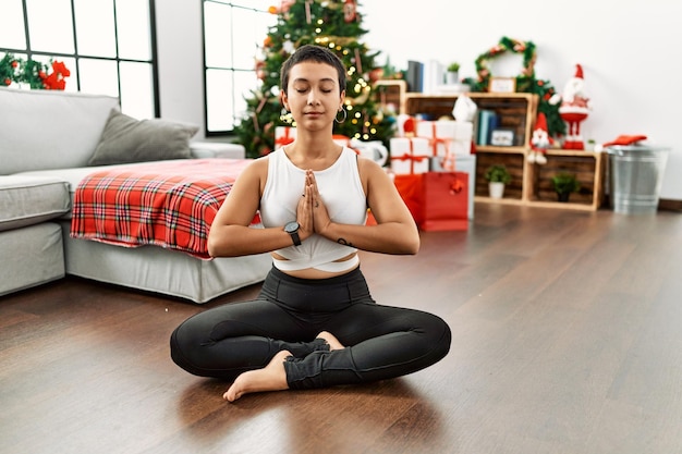 Giovane donna ispanica che fa esercizio di yoga seduto vicino all'albero di Natale a casa