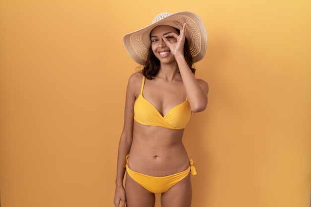 Giovane donna ispanica che indossa bikini e cappello estivo facendo un gesto ok con l'occhio sorridente della mano guardando attraverso le dita con la faccia felice