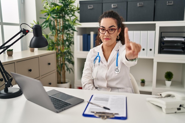 Giovane donna ispanica che indossa l'uniforme del medico e lo stetoscopio puntato con il dito verso l'alto e l'espressione arrabbiata che non mostra alcun gesto