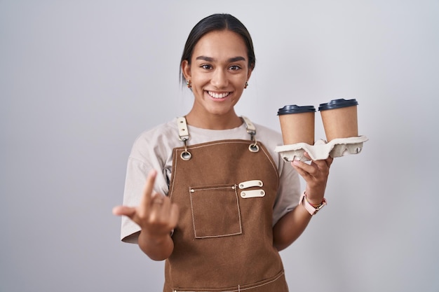 Giovane donna ispanica che indossa un grembiule da cameriera professionale che tiene il caffè che fa cenno vieni qui gesto con la mano che invita ad accogliere felice e sorridente