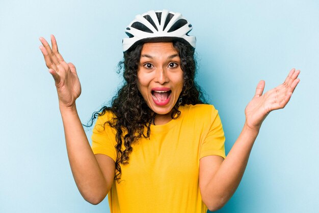 Giovane donna ispanica che indossa una bici da casco isolata su sfondo blu ricevendo una piacevole sorpresa eccitata e alzando le mani