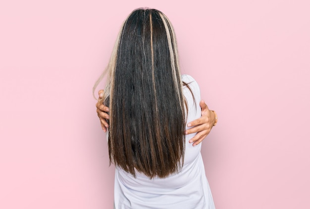 Giovane donna ispanica che indossa una maglietta bianca casual che si abbraccia felice e positiva dall'amore di sé all'indietro e dalla cura di sé