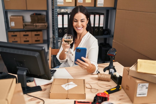 Giovane donna ispanica che lavora bevendo vino e utilizza lo smartphone sorridendo con un sorriso felice e fresco sul viso che mostra i denti