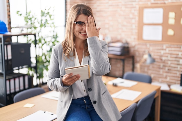 Giovane donna ispanica che lavora in ufficio con gli occhiali che coprono un occhio con la mano un sorriso sicuro sul viso e un'emozione di sorpresa