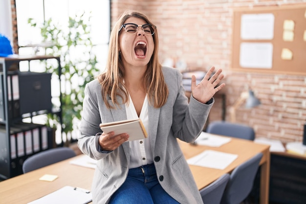 Giovane donna ispanica che lavora in ufficio con gli occhiali che gridano e urlano forte a fianco con la mano sulla bocca. concetto di comunicazione.