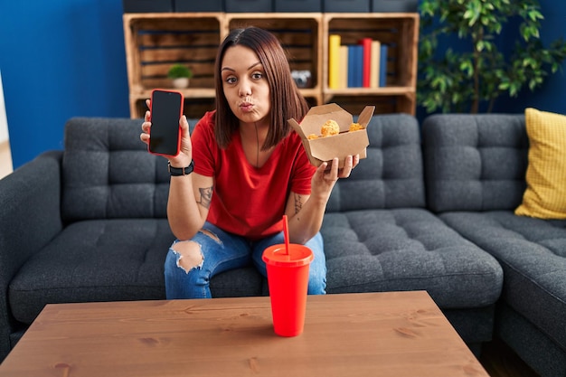 Giovane donna ispanica che mangia fast food mostrando lo schermo dello smartphone guardando la telecamera che soffia un bacio essendo adorabile e sexy. espressione d'amore.