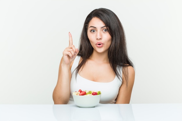 Giovane donna ispanica che mangia una ciotola di frutta che ha una certa grande idea, concetto di creatività.