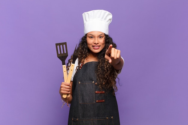 Giovane donna ispanica che punta alla telecamera con un sorriso soddisfatto, fiducioso e amichevole, scegliendo te. concetto di chef barbecue