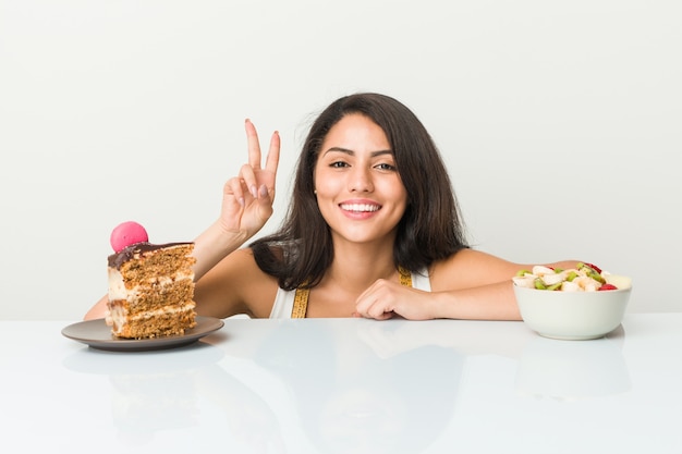 Giovane donna ispanica che sceglie fra il dolce o la frutta che mostra il segno di vittoria e che sorride largamente.