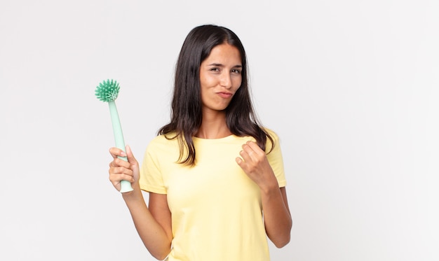 Giovane donna ispanica che sembra arrogante, di successo, positiva e orgogliosa e tiene in mano una spazzola per la pulizia dei piatti