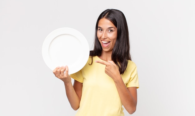 Giovane donna ispanica che sembra eccitata e sorpresa indicando il lato e tenendo in mano un piatto vuoto