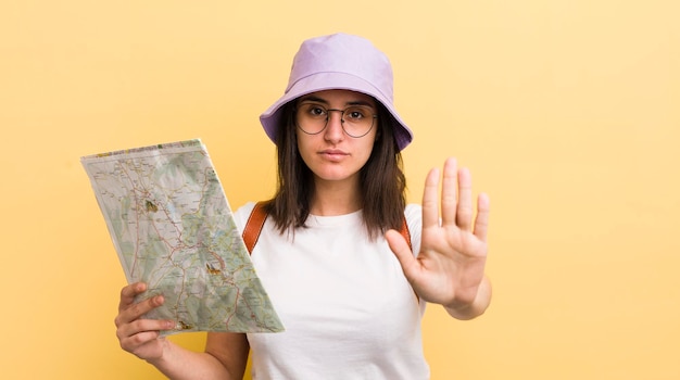 Giovane donna ispanica che sembra seria mostrando il palmo aperto facendo il gesto di arresto turistico e il concetto di mappa