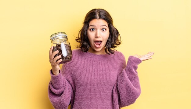 Giovane donna ispanica che sembra sorpresa e scioccata, con la mascella caduta in possesso di un oggetto. concetto di chicchi di caffè