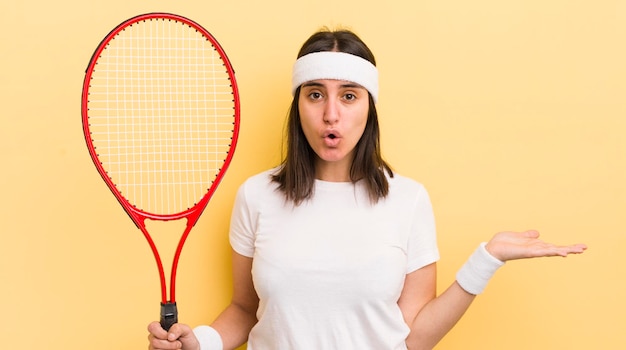 Giovane donna ispanica che sembra sorpresa e scioccata con la mascella caduta tenendo un concetto di tennis oggetto