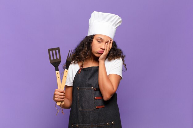 Giovane donna ispanica che si sente annoiata, frustrata e assonnata dopo un compito noioso, noioso e noioso, tenendo il viso con la mano. concetto di chef barbecue