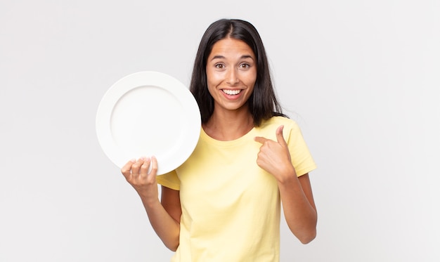 Giovane donna ispanica che si sente felice e indica se stessa con un eccitato e tiene in mano un piatto vuoto