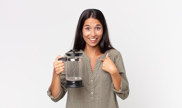 giovane donna ispanica che si sente felice e indica se stessa con un eccitato e tiene in mano una caffettiera manuale