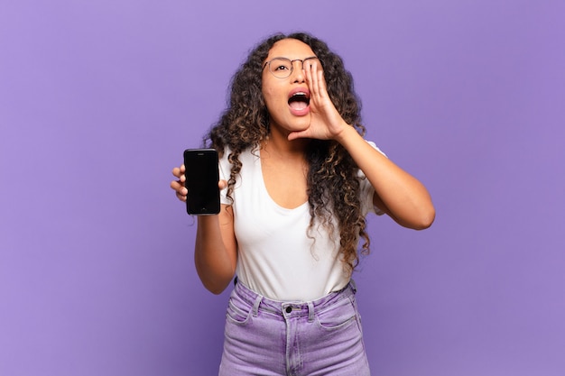 Giovane donna ispanica che si sente felice, eccitata e positiva, dando un grande grido con le mani vicino alla bocca, chiamando. concetto di smartphone