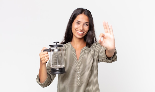 Giovane donna ispanica che si sente felice, mostra approvazione con un gesto ok e tiene in mano una caffettiera manuale