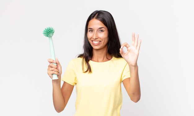 Giovane donna ispanica che si sente felice, mostra approvazione con un gesto ok e tiene in mano una spazzola per la pulizia dei piatti