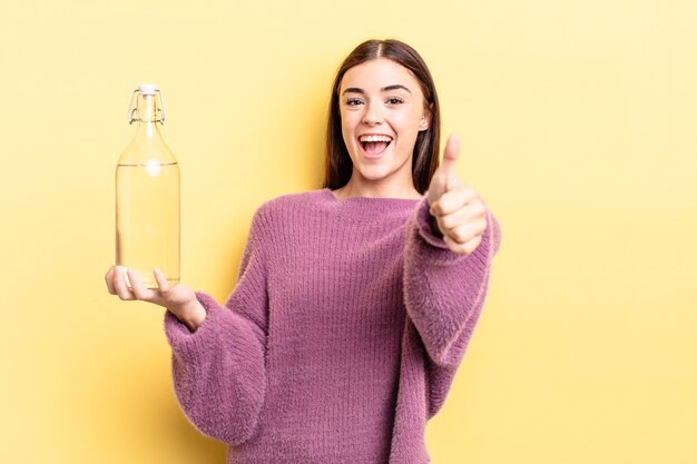 Giovane donna ispanica che si sente orgogliosa, sorride positivamente con i pollici in su. concetto di bottiglia d'acqua