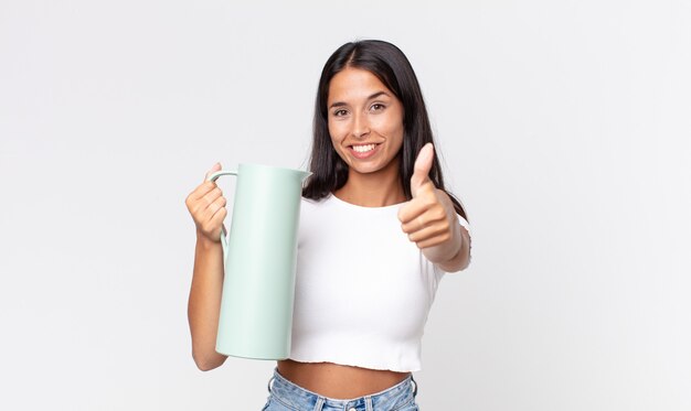 Giovane donna ispanica che si sente orgogliosa, sorride positivamente con il pollice in alto e tiene in mano un thermos di caffè