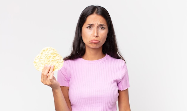 Giovane donna ispanica che si sente triste e piagnucolona con uno sguardo infelice e piange e tiene in mano un biscotto di riso. concetto di dieta