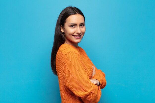 Giovane donna ispanica che sorride alla macchina fotografica con le braccia incrociate e un'espressione felice, fiduciosa, soddisfatta, vista laterale