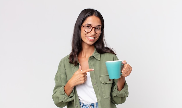 Giovane donna ispanica che sorride allegramente, si sente felice e indica il lato e tiene in mano una tazza da caffè