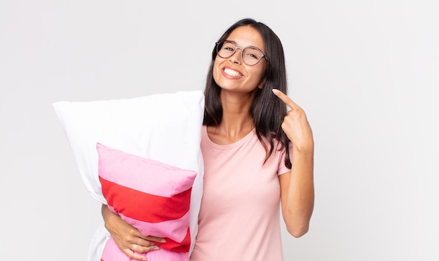 Giovane donna ispanica che sorride con sicurezza indicando il proprio ampio sorriso che indossa un pigiama e tiene in mano un cuscino