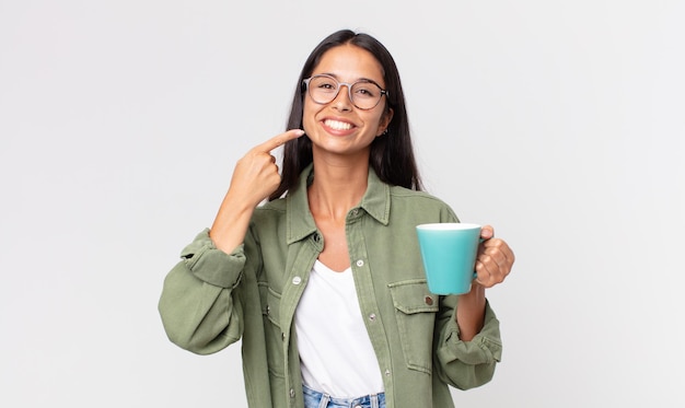 Giovane donna ispanica che sorride con sicurezza indicando il proprio ampio sorriso e tenendo in mano una tazza da caffè