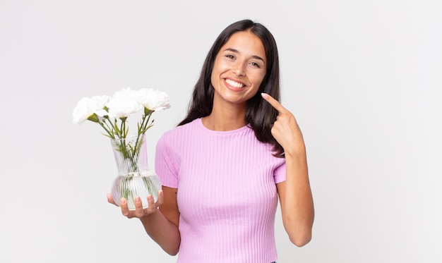 giovane donna ispanica che sorride con sicurezza indicando il proprio ampio sorriso tenendo fiori decorativi