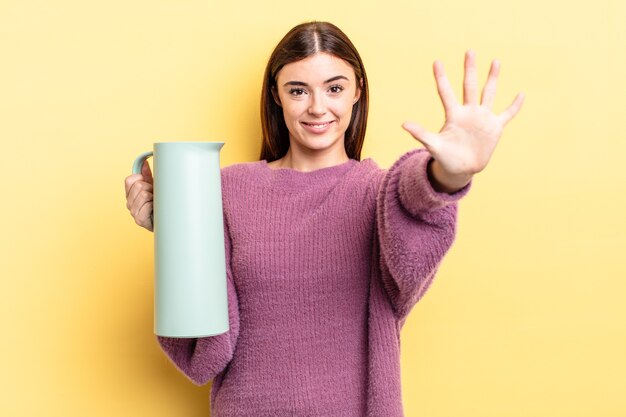 Giovane donna ispanica che sorride e che sembra amichevole, mostrando il numero cinque. concetto di thermos per caffè