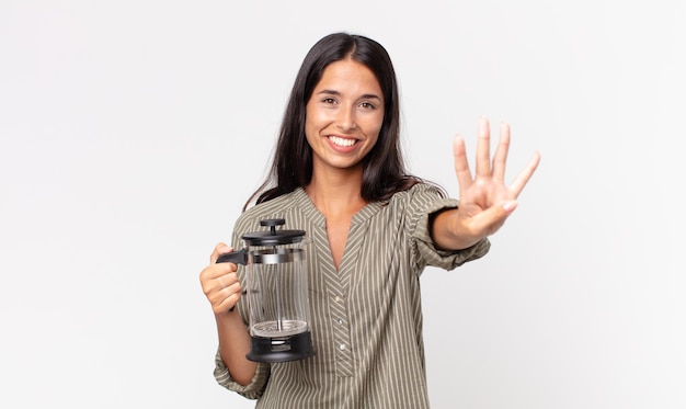 Giovane donna ispanica che sorride e sembra amichevole, mostra il numero quattro e tiene in mano una caffettiera manuale